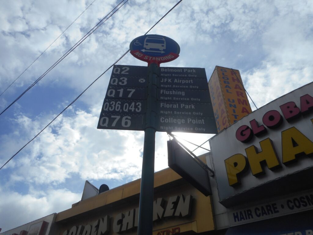 City bus stop sign with routes, destinations, and nearby commercial businesses in an urban setting.