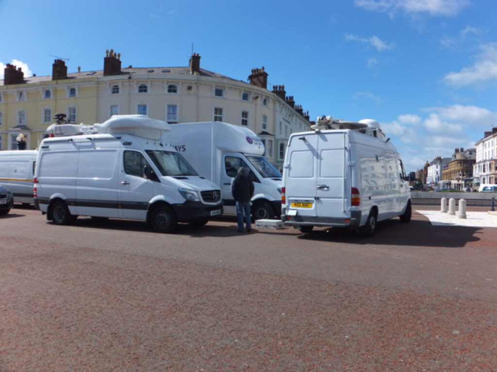 Media vans with satellite dishes parked near historic buildings in a vibrant urban streetscape.