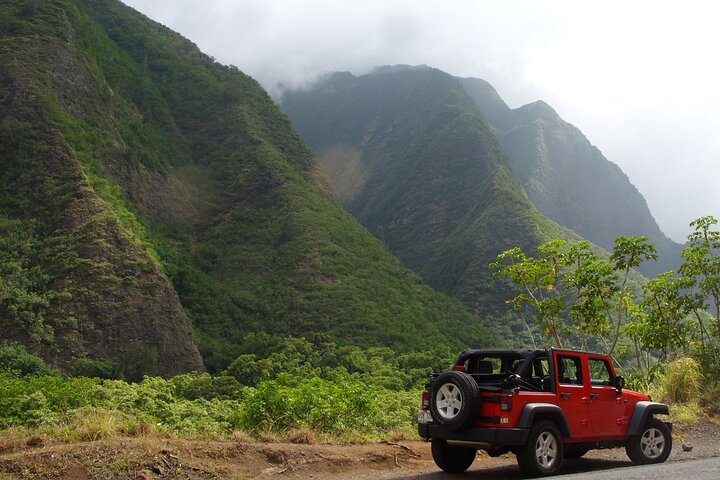 Red Jeep parked near lush green mountains under cloudy sky, perfect for adventure.