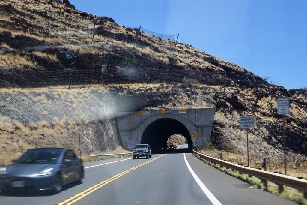 Mountain tunnel highway scene with vehicles, rocky terrain, and clear blue sky above.