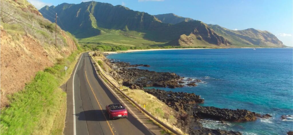 Scenic coastal drive features a red convertible, ocean views, and majestic mountains.